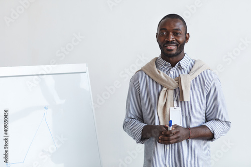 Waist up portrait of African-American business coach talking to audience at conference or education seminar while standing by whiteboard and smiling, copy space
