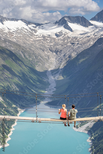 Young couple sit on swing bridge facing on reservoir glacier in Zillertal Austria
