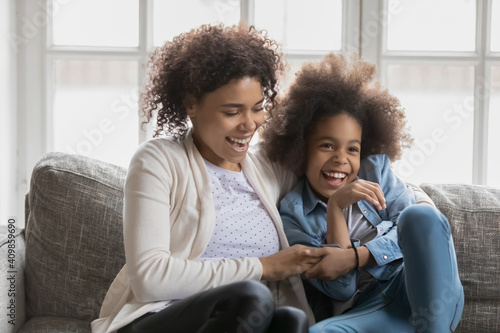 Close up overjoyed African American mother and daughter tickling, hugging, sitting on cozy couch at home, happy mum and little adorable girl child having fun, enjoying leisure time together