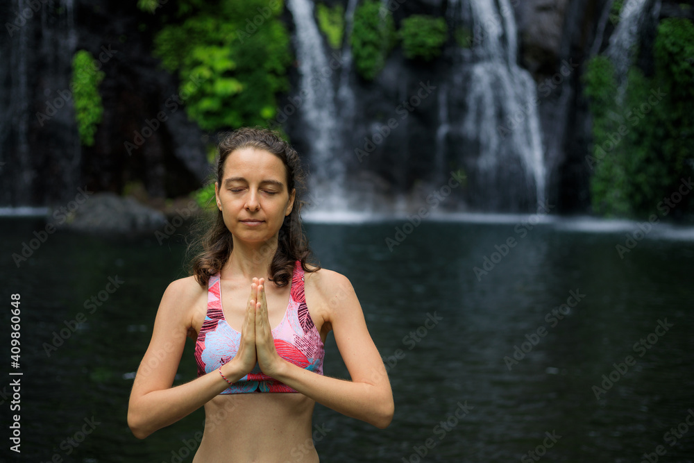Hands raising up in namaste mudra. Yoga near waterfall. Young woman meditating, practicing yoga and pranayama. Copy space. View from back. Yoga retreat. Banyumala waterfall, Bali