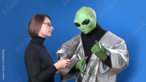 Woman journalist in glasses with microphone interviews green skinned alien in silver clothes on blue chromakey background photo