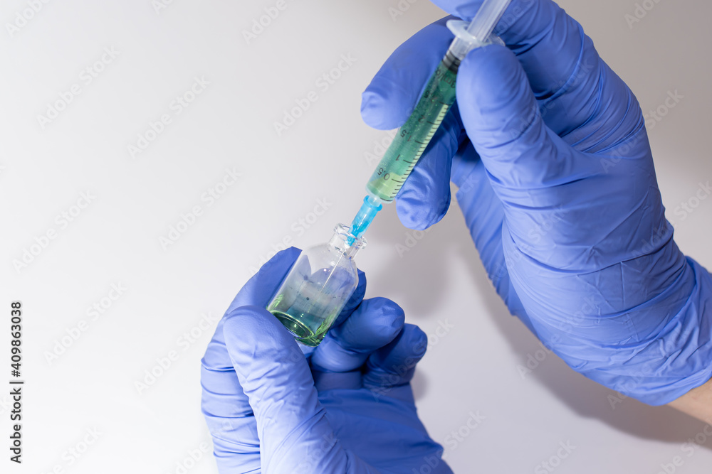 Hands in medical gloves hold a bottle with a vaccine on a white background. The syringe is filled with a liquid containing a green medicine. Hands in medical gloves hold a bottle with a vaccine on a white background. The syringe is filled with a liquid containing a green medicine.