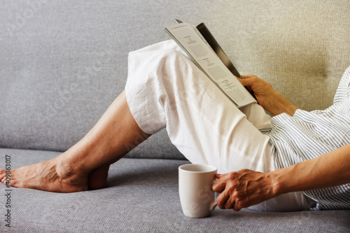 Middle-aged brunette woman with glasses on the gray sofa reading magazine with cup of coffee, soft focus