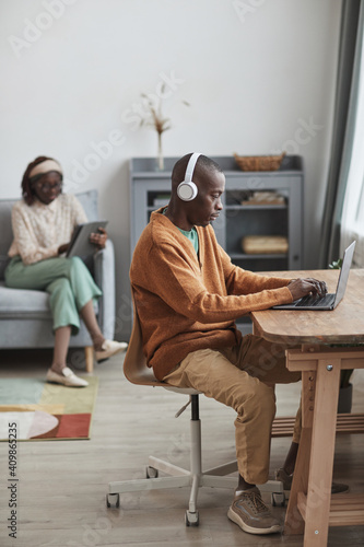 Vertical portrait of contemporary couple using internet devices at home, focus on African-American man wearing headphones and typing at desk in foreground, copy space