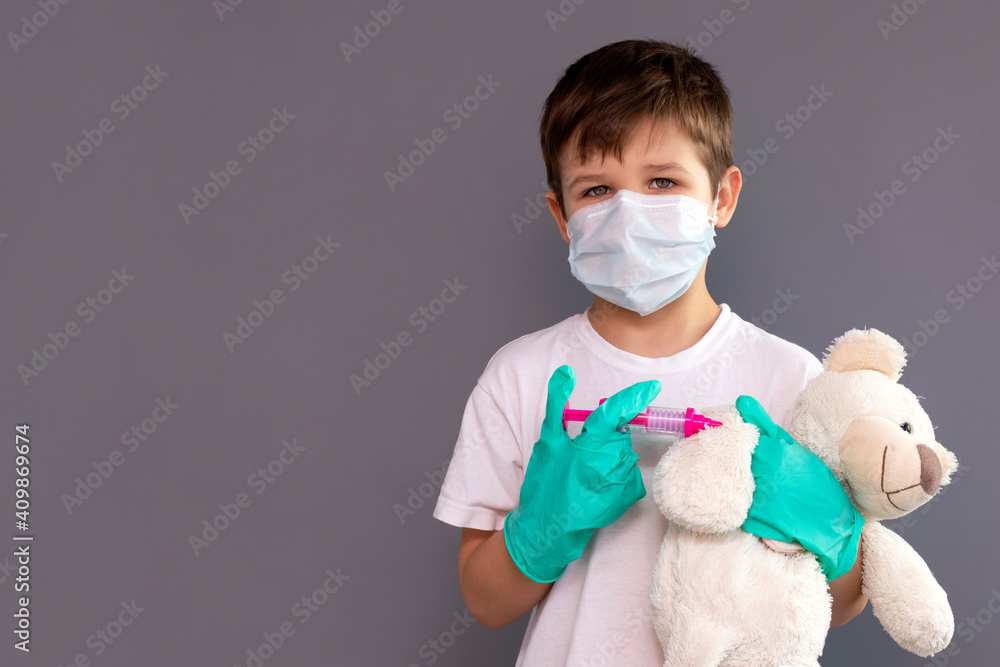 A little boy in a medical mask holds a toy syringe and a teddy bear in his hand. Game concept. Vaccination theme. Healthcare and medicine. Covid 19