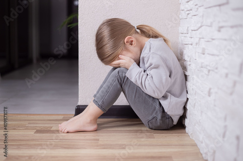 Sad little child girl sitting on floor in corner at home.