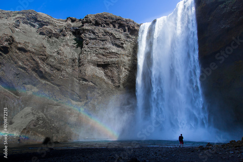 Horizontal view of the renowned Skógafoss waterfall with a double rainbow seen during a sunny spring morning, with man standing at its foot, Skógá, southern Iceland