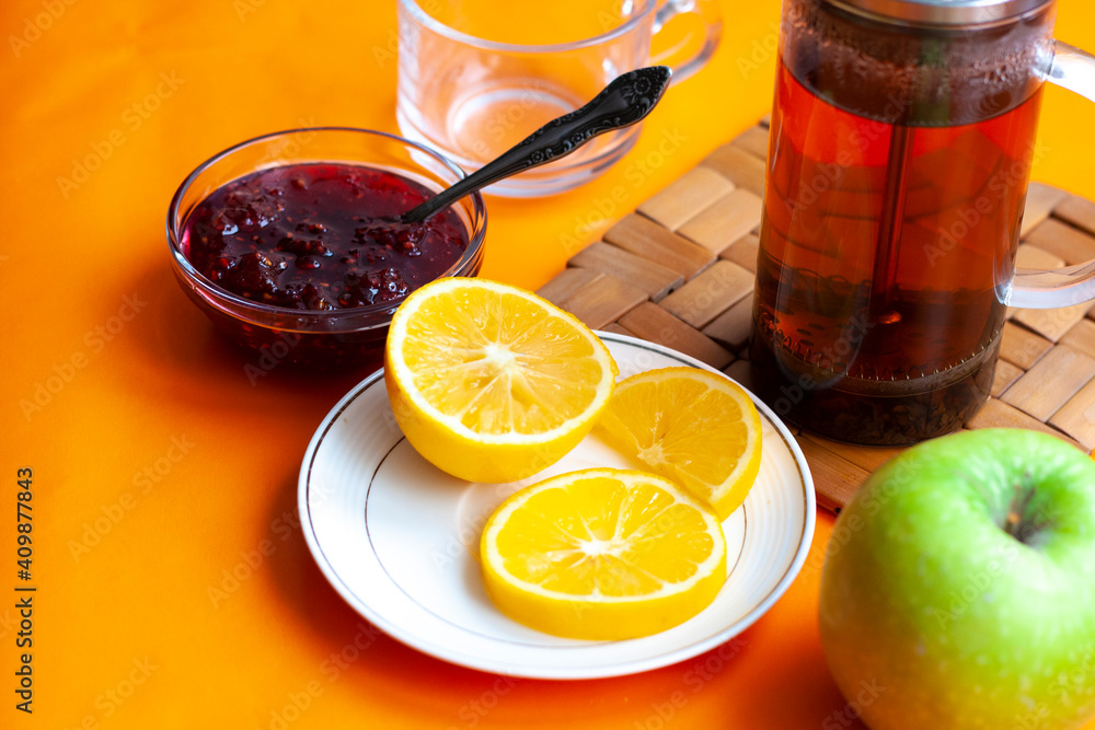 Breakfast tea in French press, lemon, cup, jam with spoon, apple, orange background Breakfast tea in French press, lemon, cup, jam with spoon, apple, orange background
