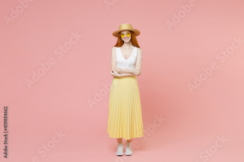 Full length body young redhead ginger woman 20s wearing straw hat glasses summer clothes yellow pleated skirt hold hands crossed folded look camera isolated on pastel pink background studio portrait.