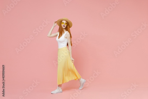 Full length body young redhead ginger woman in straw hat glasses summer clothes yellow pleated maxi skirt looking camera walking going passing isolated on pastel pink color background studio portrait.