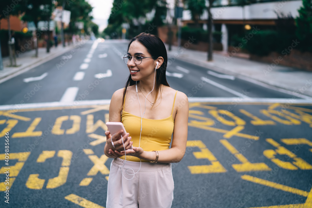 Happy hipster girl in electronic headphones enjoying favorite music standing at urban road without traffic, smiling female millennial listening audio playlist from modern smartphone device