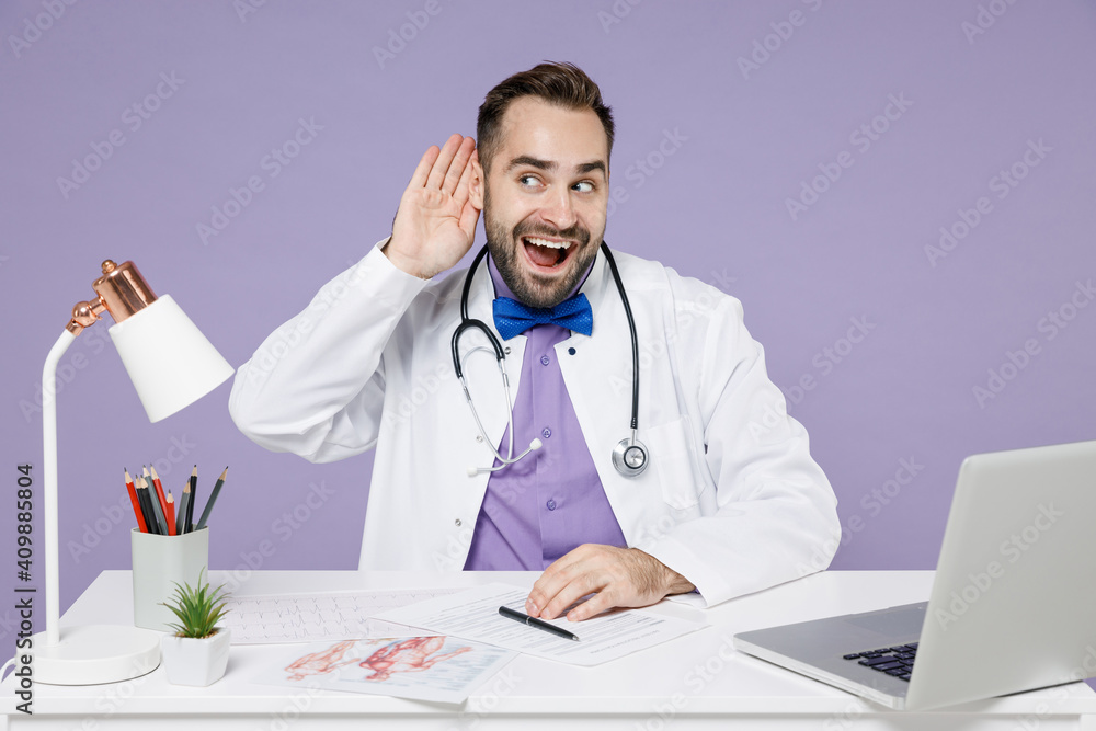 Male doctor man in white medical gown suit sits at desk work on computer documents in hospital clinic office try to hear you overhear listening intently isolated on violet background studio portrait Male doctor man in white medical gown suit sits at desk work on computer documents in hospital clinic office try to hear you overhear listening intently isolated on violet background studio portrait