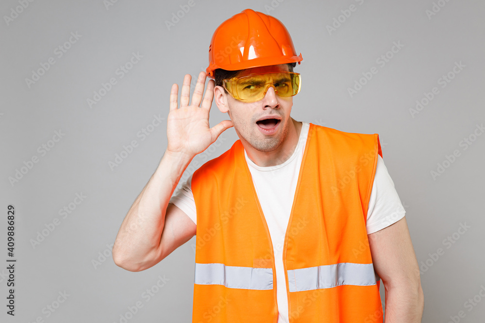Young employee man in orange vest protective helmet hardhat try hear you overhear listening intently isolated on grey background studio. Instruments for renovation apartment room. Repair home concept. Young employee man in orange vest protective helmet hardhat try hear you overhear listening intently isolated on grey background studio. Instruments for renovation apartment room. Repair home concept.