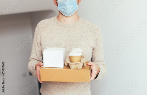 A delivery man in medical mask with cardboard brown and white boxes and take away coffee cups delivering breakfast to a customer's home