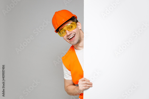 Young employee handyman man in orange vest protective hardhat hold blank sign board with place for text isolated on grey background studio. Instruments for renovation apartment. Repair home concept. photo