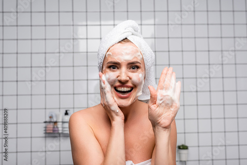 cheerful woman with naked shoulders showing palm with foam cleanser in bathroom photo