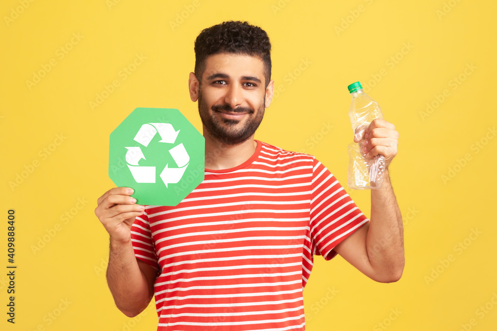 Happy satisfied man with beard in striped t-shirt pressing bottle holding in hand recycling sign board, plastic disposal, garbage sorting. Indoor studio shot isolated on yellow background Happy satisfied man with beard in striped t-shirt pressing bottle holding in hand recycling sign board, plastic disposal, garbage sorting. Indoor studio shot isolated on yellow background