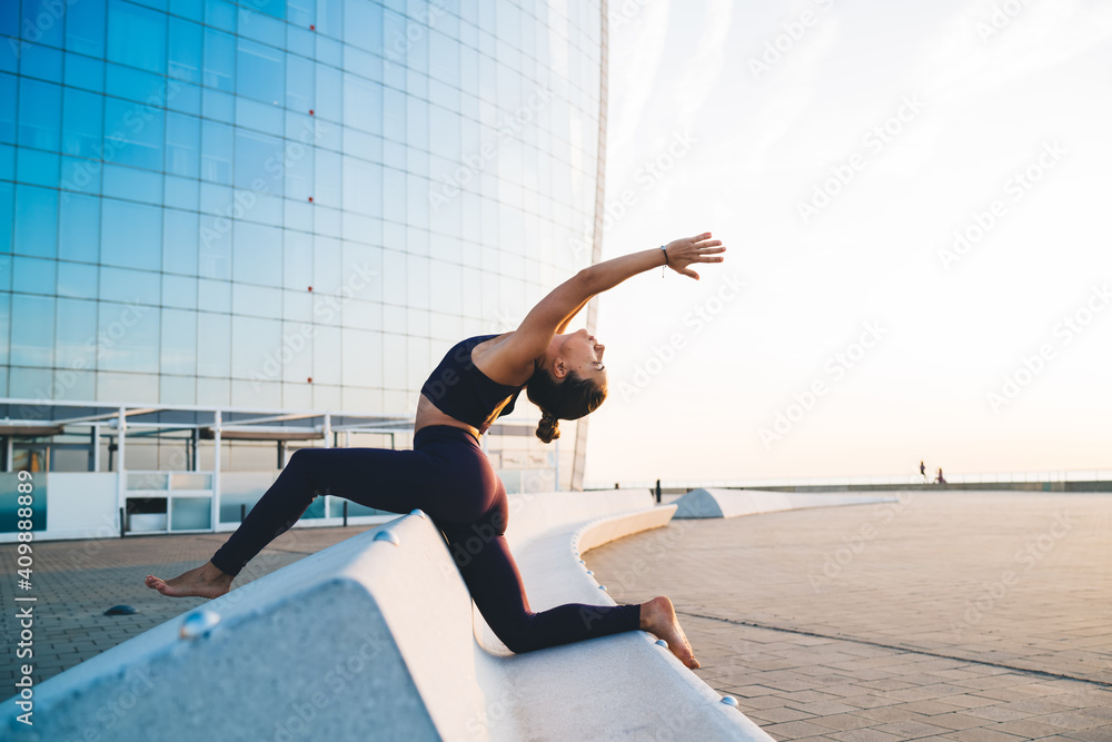 Experienced sportswoman with perfect body shape practicing pilates hatha yoga during leisure in city, inspired female enjoying morning workout for training stretching standing in Crescent pose