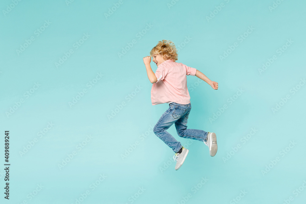 Full length side view of funny little kid boy 10s in casual pink shirt jumping doing winner gesture isolated on blue turquoise color background children studio portrait. Childhood lifestyle concept. Full length side view of funny little kid boy 10s in casual pink shirt jumping doing winner gesture isolated on blue turquoise color background children studio portrait. Childhood lifestyle concept.