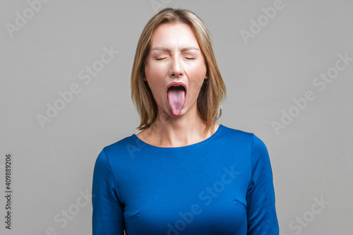 Portrait of positive carefree woman with fair hair in blue dress standing with closed eyes and demonstrating tongue, naughty disobedient expression. indoor studio shot isolated on gray background