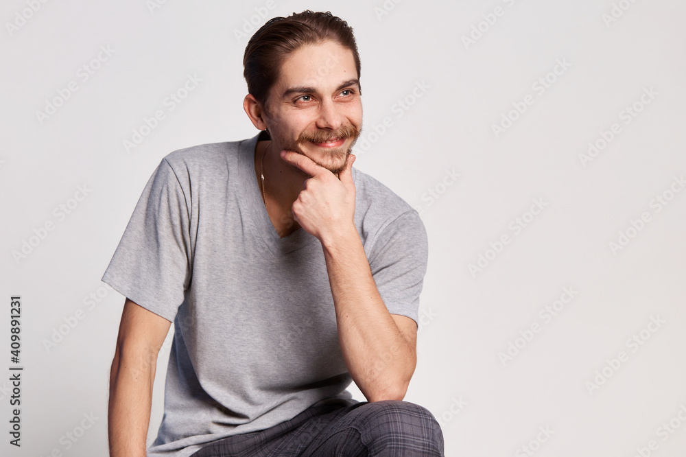 Handsome young man in shirt sitting on the chair with a grin and and holds his hand near his face isolated on a gray background Handsome young man in shirt sitting on the chair with a grin and and holds his hand near his face isolated on a gray background
