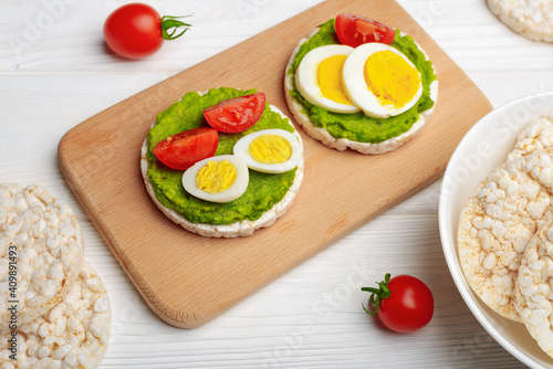 Rice cakes with avocado mash and eggs with tomato on cutting board and white wooden background