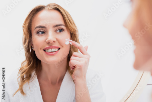 reflection of smiling woman applying cosmetic cream on face in bathroom