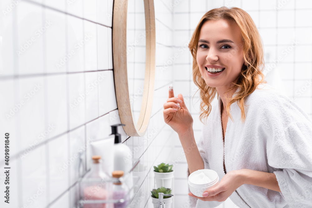 smiling woman holding jar with face cream near mirror near bottles on blurred foreground smiling woman holding jar with face cream near mirror near bottles on blurred foreground