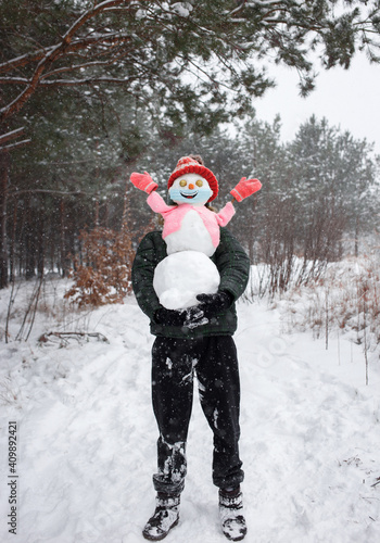 unrecognizable figure of man in snow holds in arms cheerful snowman in red knitted hat, jacket, mittens, medical mask with smile painted on it. Winter fun, healthy lifestyle, digital detox photo