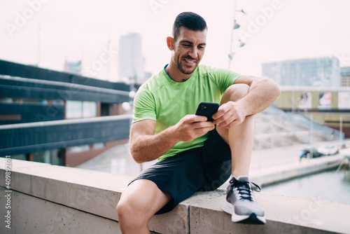 Happy male runner enjoying cellular messaging with personal trainer resting at urbanity after morning cardio workout, cheerful muscular sportsman using 4g wireless internet for sending publication