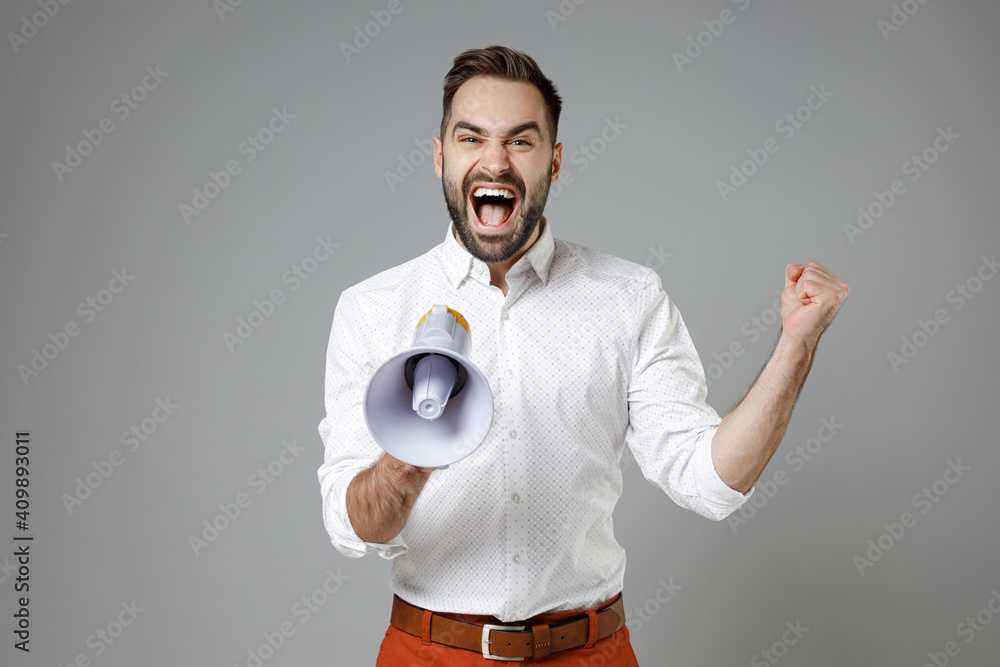 Overjoyed young bearded business man in classic white shirt screaming in megaphone doing winner gesture clenching fist isolated on grey background studio portrait. Achievement career wealth concept. Overjoyed young bearded business man in classic white shirt screaming in megaphone doing winner gesture clenching fist isolated on grey background studio portrait. Achievement career wealth concept.