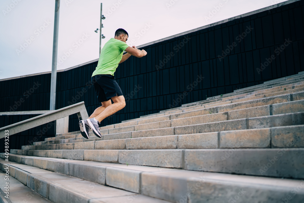 Back view of muscular male bodybuilder dressed in sportive clothing jumping at urban stairs getting high with effort, determined male runner warming up body during cardio training for losing weight