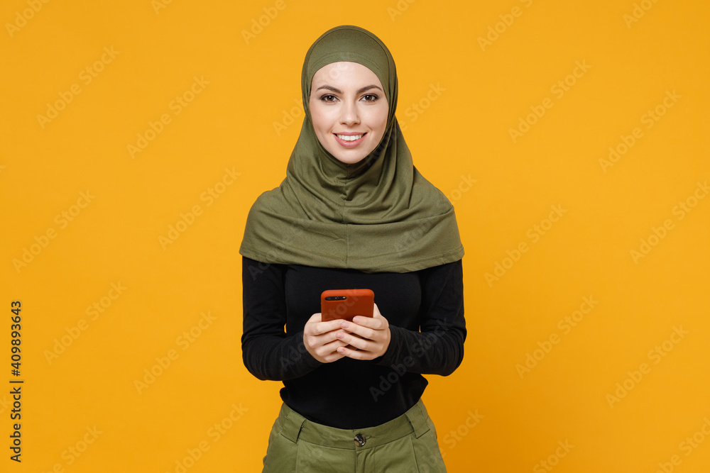 Smiling young arabian muslim woman wearing hijab black green clothes using mobile cell phone typing sms message isolated on yellow color background studio portrait. People religious lifestyle concept. Smiling young arabian muslim woman wearing hijab black green clothes using mobile cell phone typing sms message isolated on yellow color background studio portrait. People religious lifestyle concept.