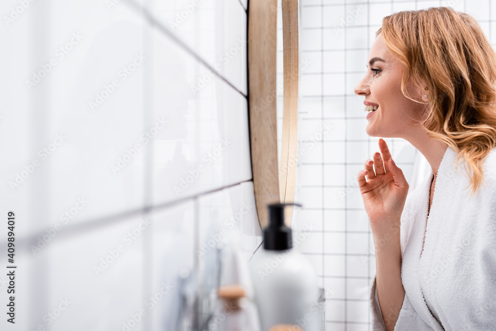 side view of happy woman looking at mirror near bottles on blurred foreground side view of happy woman looking at mirror near bottles on blurred foreground