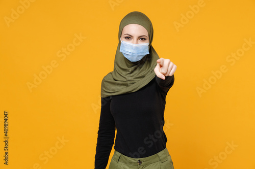 Arabian muslim woman in hijab green clothes sterile face mask to safe from coronavirus virus covid-19 pointing index finger on camera isolated on yellow background. People religious lifestyle concept.