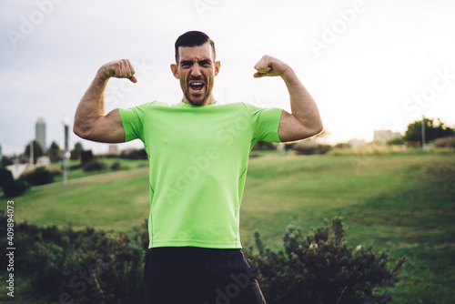 Half length portrait of muscular athletic man showing pumped up arms biceps during cardio training in park, Caucasian bodybuilder feeling confidence and power strength proud of own sportive goals
