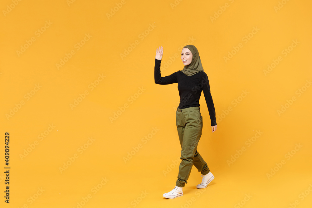 Full length side view funny young arabian muslim woman in hijab black green clothes waving and greeting with hand as notices someone isolated on yellow background. People religious lifestyle concept. Full length side view funny young arabian muslim woman in hijab black green clothes waving and greeting with hand as notices someone isolated on yellow background. People religious lifestyle concept.