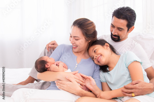 Proudly Asian man hug wife and 2 kids with love, care, protection, adorable daughter lie down on father's bosom or chest looking newborn brother in mother hand play infant feet, happy family concept