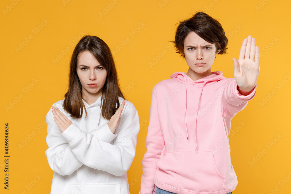 Dissatisfied two young women friends 20s wearing casual basic white pink hoodies standing holding palm folded crossed hands in stop gesture isolated on bright yellow color background studio portrait. Dissatisfied two young women friends 20s wearing casual basic white pink hoodies standing holding palm folded crossed hands in stop gesture isolated on bright yellow color background studio portrait.