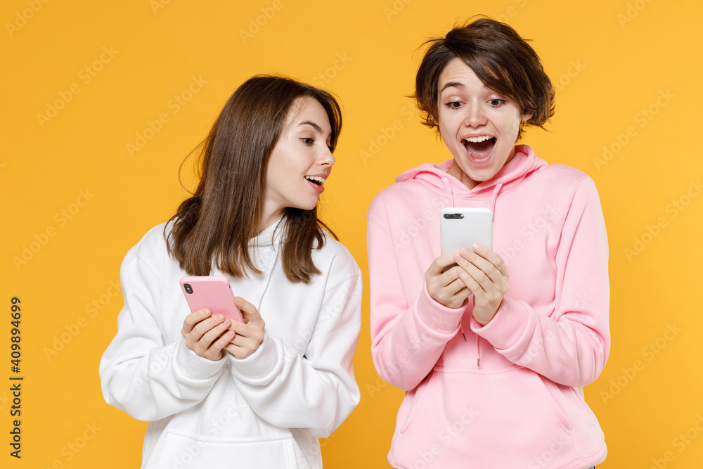 Excited amazed smiling two young women friends 20s wearing casual white pink hoodies standing using mobile cell phone typing sms message isolated on bright yellow color background studio portrait. Excited amazed smiling two young women friends 20s wearing casual white pink hoodies standing using mobile cell phone typing sms message isolated on bright yellow color background studio portrait.