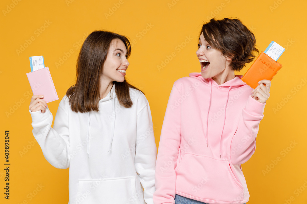 Excited cheerful two young traveler tourist women friends 20s in casual white pink hoodies hold passport tickets boarding pass looking at each other isolated on yellow background studio portrait. Excited cheerful two young traveler tourist women friends 20s in casual white pink hoodies hold passport tickets boarding pass looking at each other isolated on yellow background studio portrait.
