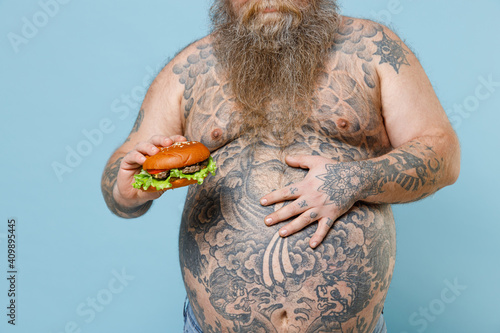 Cropped image of fat pudge obese chubby overweight man has tattooed naked big belly hold fast food burger put hand on stomach isolated on blue background. Weight loss obesity unhealthy diet concept.