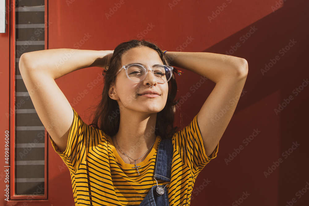 © EZ PHOTOS - Portrait of a beautiful caucasian teenager girl wearing transparent trendy glasses and colorful clothes with her hands behind her head smiling in relief. Dark red background.