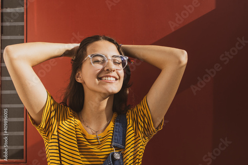 Portrait of a beautiful caucasian teenager girl wearing transparent trendy glasses and colorful clothes with her hands behind her head smiling with eyes shut in relief. Dark red background.  photo