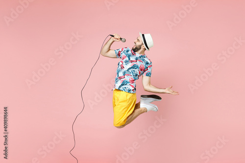 Full length side view cheerful young traveler tourist man in summer clothes hat jumping sing song in microphone isolated on pink background. Passenger traveling on weekend. Air flight journey concept.