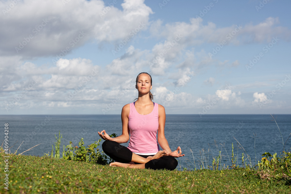 young beautiful smiling blonde woman in sportswear doing yoga sitting on the grass with blue sea and sky on the background