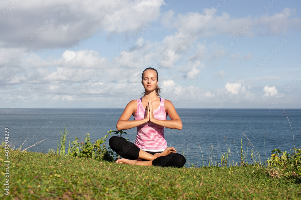 attractive smiling blonde girl in sportswear doing yoga sitting on the grass with blue sea and sky on the background