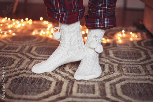 Legs in red check pyjama pants, white warm socks with pom-poms on a carpet, cosy Christmas lights on the background