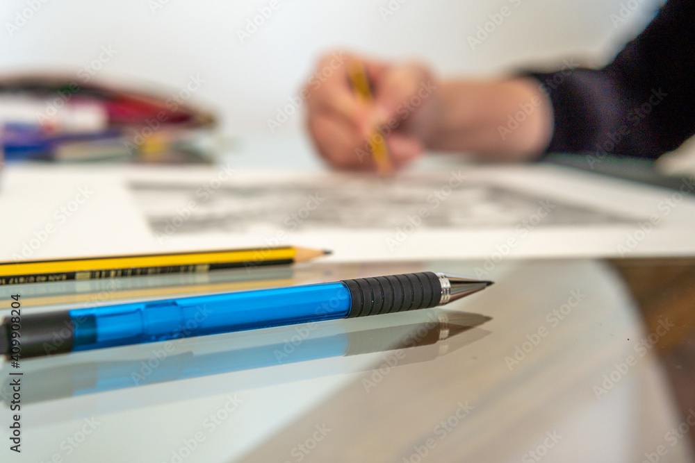 Close-up of a blue pen on a glass table, in the background out of focus the hands of an illustrator making a drawing in an artist's studio Close-up of a blue pen on a glass table, in the background out of focus the hands of an illustrator making a drawing in an artist's studio