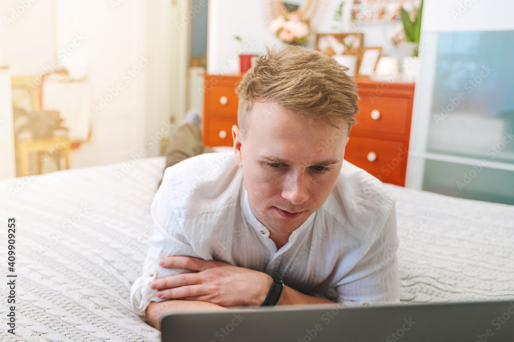 A handsome man in a shirt uses a laptop while lying on the bed. The concept of the home office and online education.